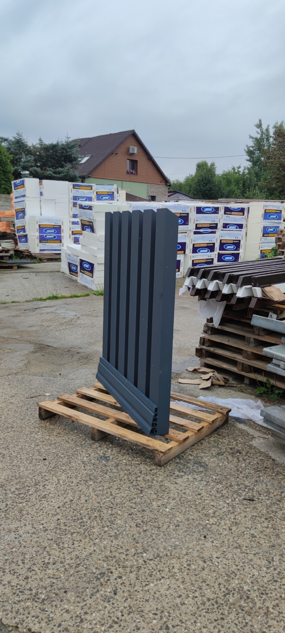Elementy ogrodzenia panelowego w kolorze grafitowym, ułożone na palecie, czekają na montaż. W tle materiały budowlane i budynek.