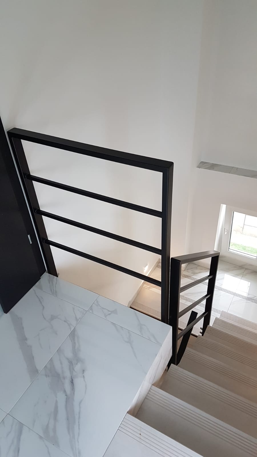 Nowoczesna, czarna balustrada schodowa z trzema poziomymi prętami, widok z góry na białe marmurowe płytki i jasne schody. Minimalistyczny design.