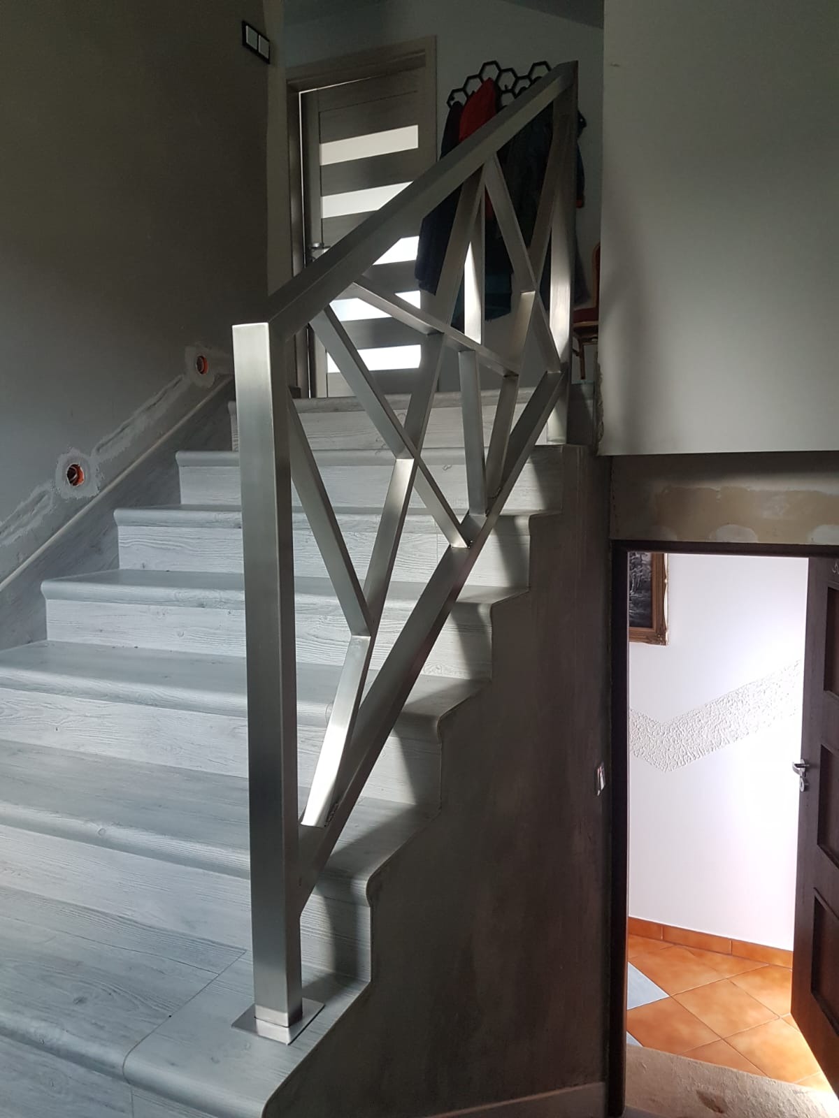 Nowoczesna balustrada schodowa ze stali nierdzewnej o geometrycznym wzorze, zamontowana na drewnianych schodach w jasnym wnętrzu domu.