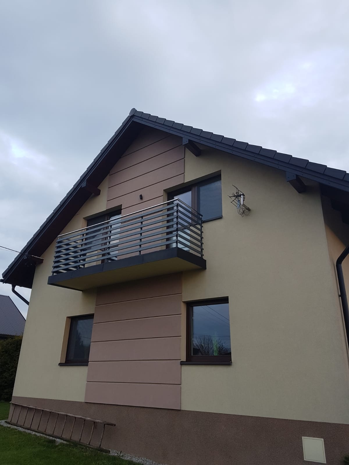 Nowoczesny balkon z metalową balustradą w domu jednorodzinnym, widok z dołu. Balustrada pozioma, szara. Na ścianie antena satelitarna. Drabina oparta o ścianę.