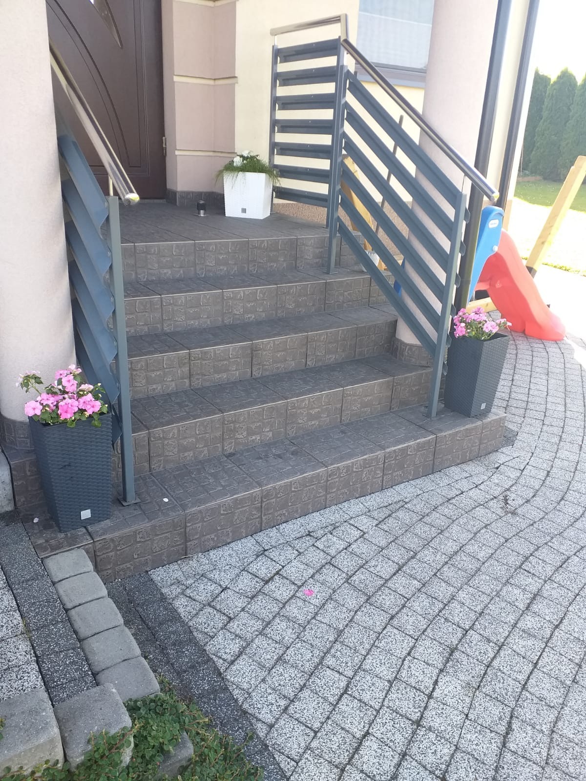 Schody wejściowe z szarą, nowoczesną balustradą metalową i donicami z kwiatami. Widoczne płytki schodowe i kostka brukowa na podjeździe.