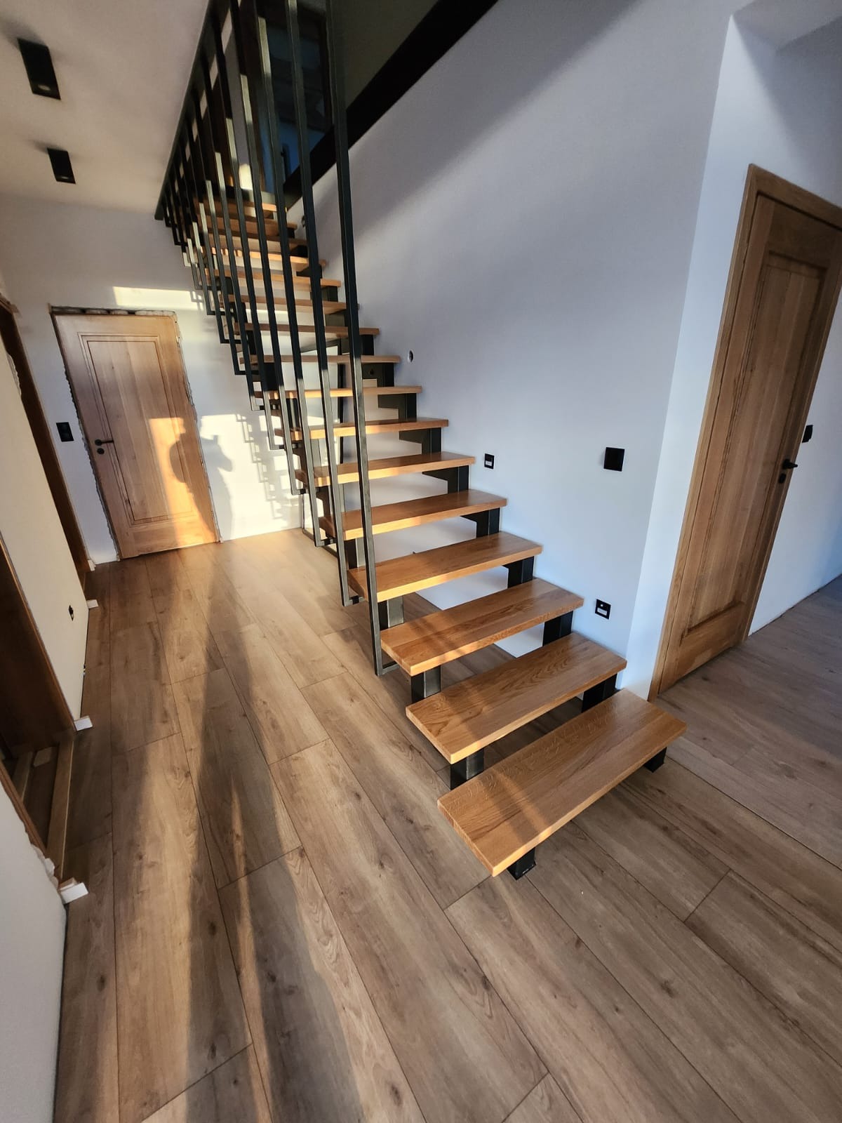 Nowoczesne schody z drewnianymi stopniami i metalową balustradą w jasnym wnętrzu. Widoczne drzwi z naturalnego drewna i podłoga w odcieniach brązu. Minimalistyczny design.
