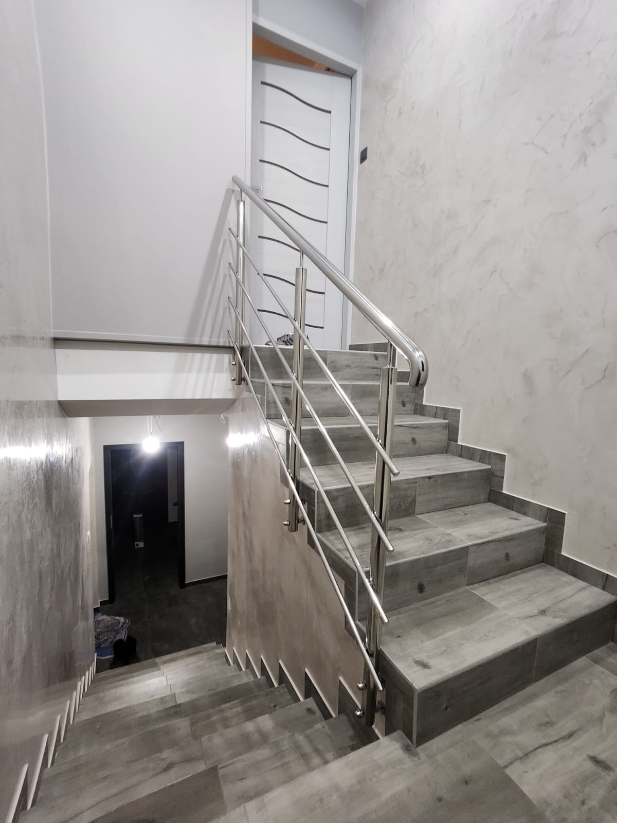 Klatka schodowa z szarymi płytkami i nowoczesną balustradą ze stali nierdzewnej. Widok z dołu, oświetlona lampą sufitową. Ściany z efektem dekoracyjnym.