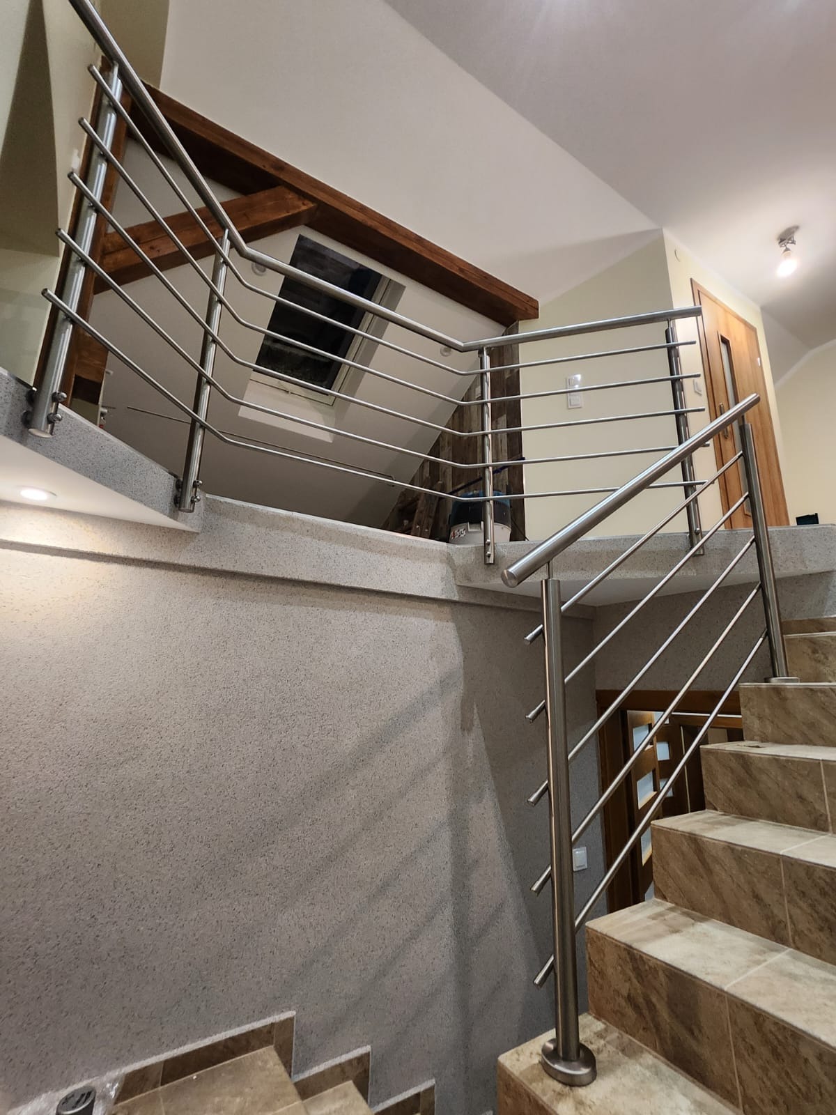 Wnętrze domu: schody z beżowych płytek z balustradą ze stali nierdzewnej. Widok z dołu na poddasze z oknem dachowym i drewnianą belką.