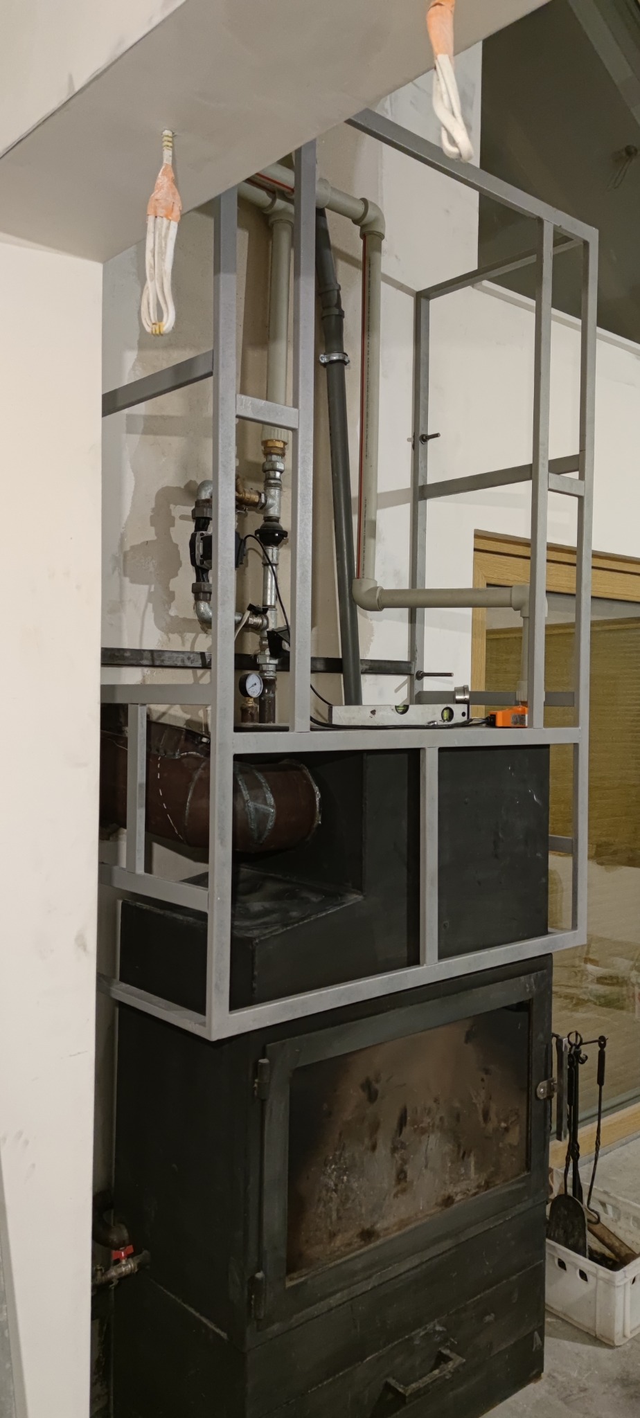 Metalowa konstrukcja obudowy kominka z widocznymi rurami instalacji, poziomica i narzędzia ślusarskie w tle. Surowy stan wykończenia wnętrza.