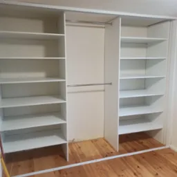 Wnętrze białej szafy wnękowej z półkami i drążkiem na ubrania, widok na drewnianą podłogę. Minimalistyczny design, surowe wykończenie.