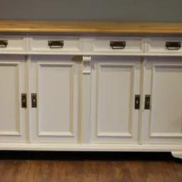 Biała komoda z czterema szufladami i drzwiczkami, blatem z jasnego drewna, metalowe uchwyty, klasyczny styl, na brązowej podłodze, szara ściana w tle.