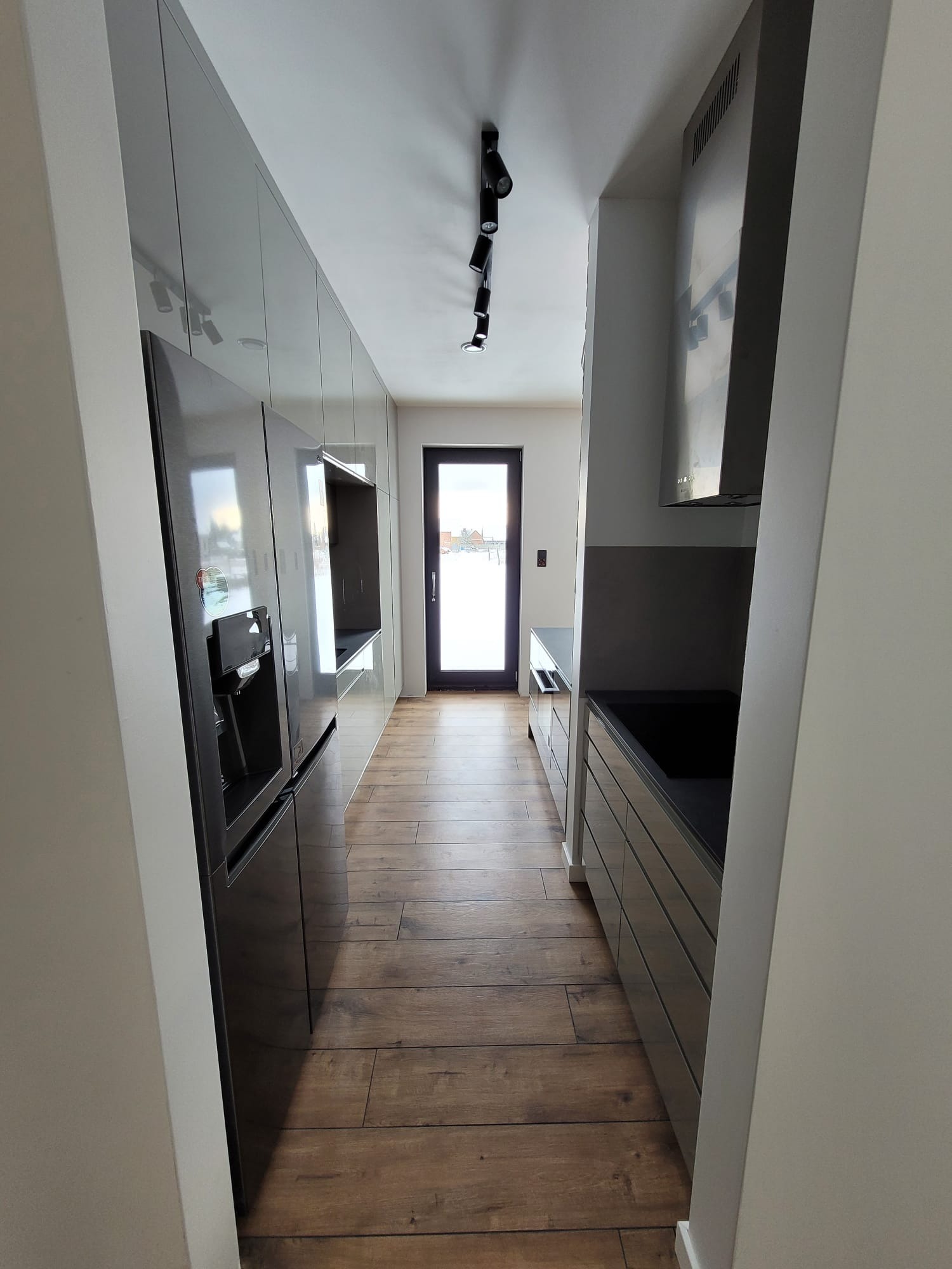 Wąskie ujęcie na nowoczesną kuchnię z błyszczącymi, szarymi frontami szafek i drewnianą podłogą. Widoczna czarna lodówka z dystrybutorem wody i czarne reflektory sufitowe.