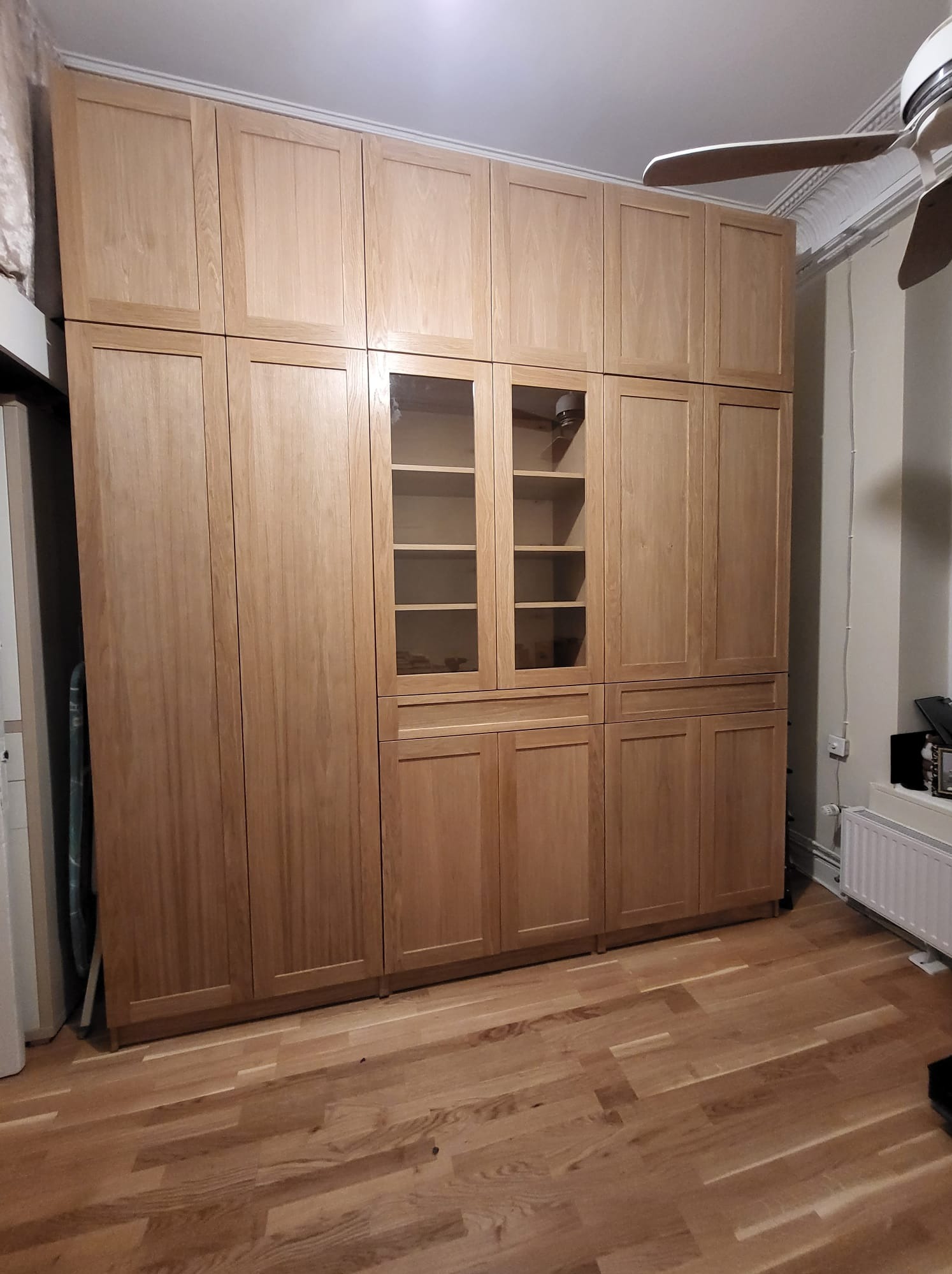 Duża, jasna szafa z drewna z przeszklonymi elementami, zajmująca całą ścianę, dopasowana do podłogi w ciepłym odcieniu brązu. Widoczny fragment sufitu z wentylatorem.