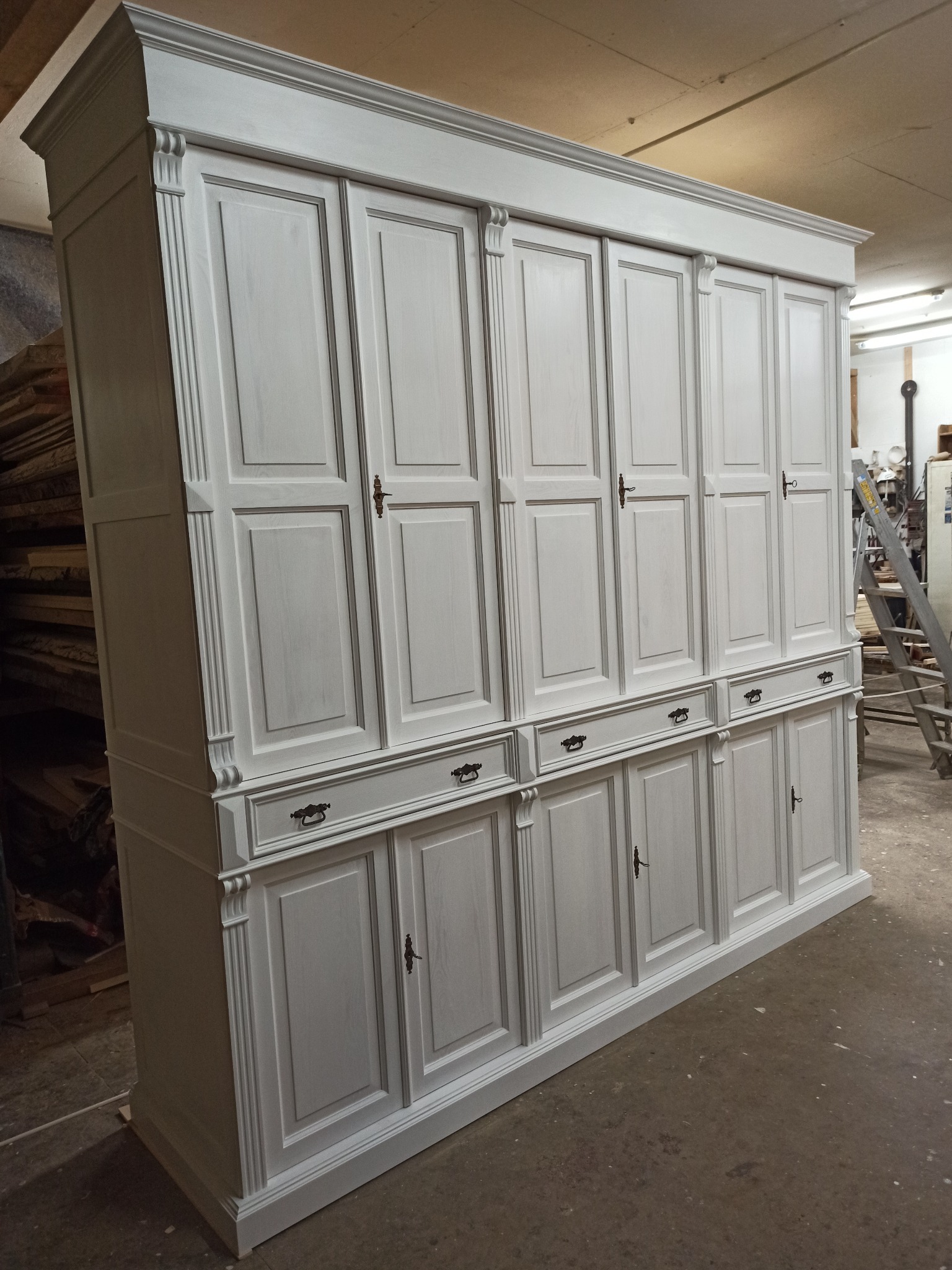 Biała, duża szafa z szufladami i zdobieniami, widoczna z boku na tle warsztatu. Elementy dekoracyjne na górze i po bokach. Metalowe uchwyty.