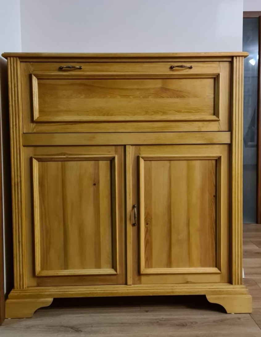 Drewniana komoda z dwoma drzwiczkami i szufladą u góry, wykonana w klasycznym stylu. Widoczne detale wykończenia i naturalna faktura drewna.