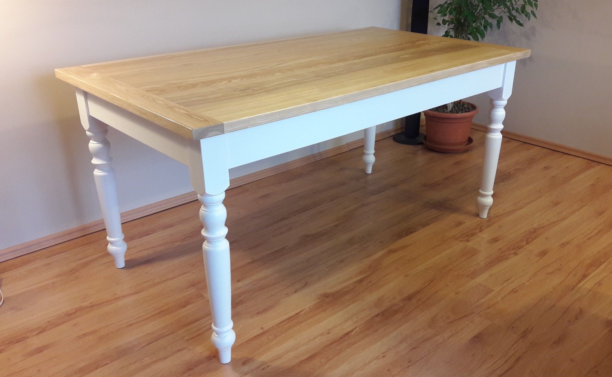 Drewniany stół z blatem w odcieniu jasnego dębu i białymi, toczonymi nogami. Klasyczny design, widoczne usłojenie drewna, ciepłe oświetlenie. Na podłodze panele.