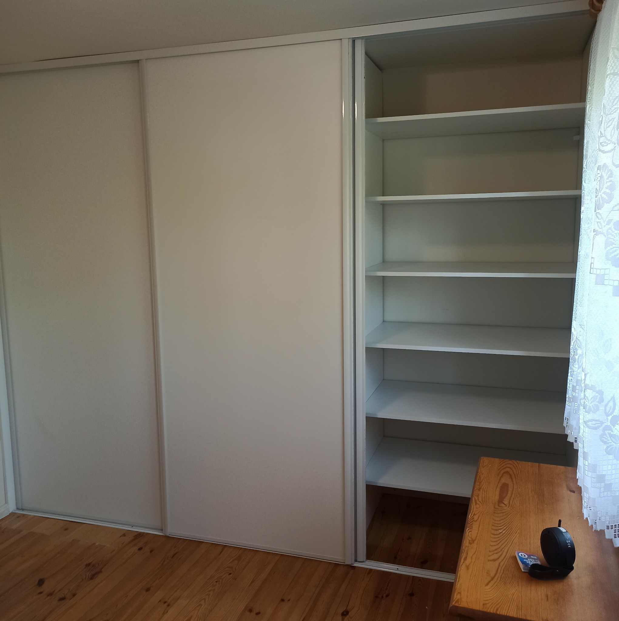 Biała szafa wnękowa z przesuwnymi drzwiami, częściowo otwarta, ukazująca puste półki. Widoczna drewniana podłoga i fragment stolika z przedmiotami.