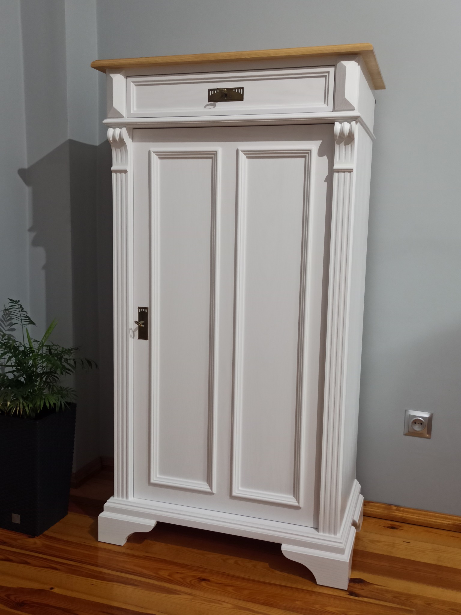 Biała szafa z szufladą i drewnianym blatem w stylu retro, widoczna roślina w czarnej donicy. Realizacja z Pomorska.