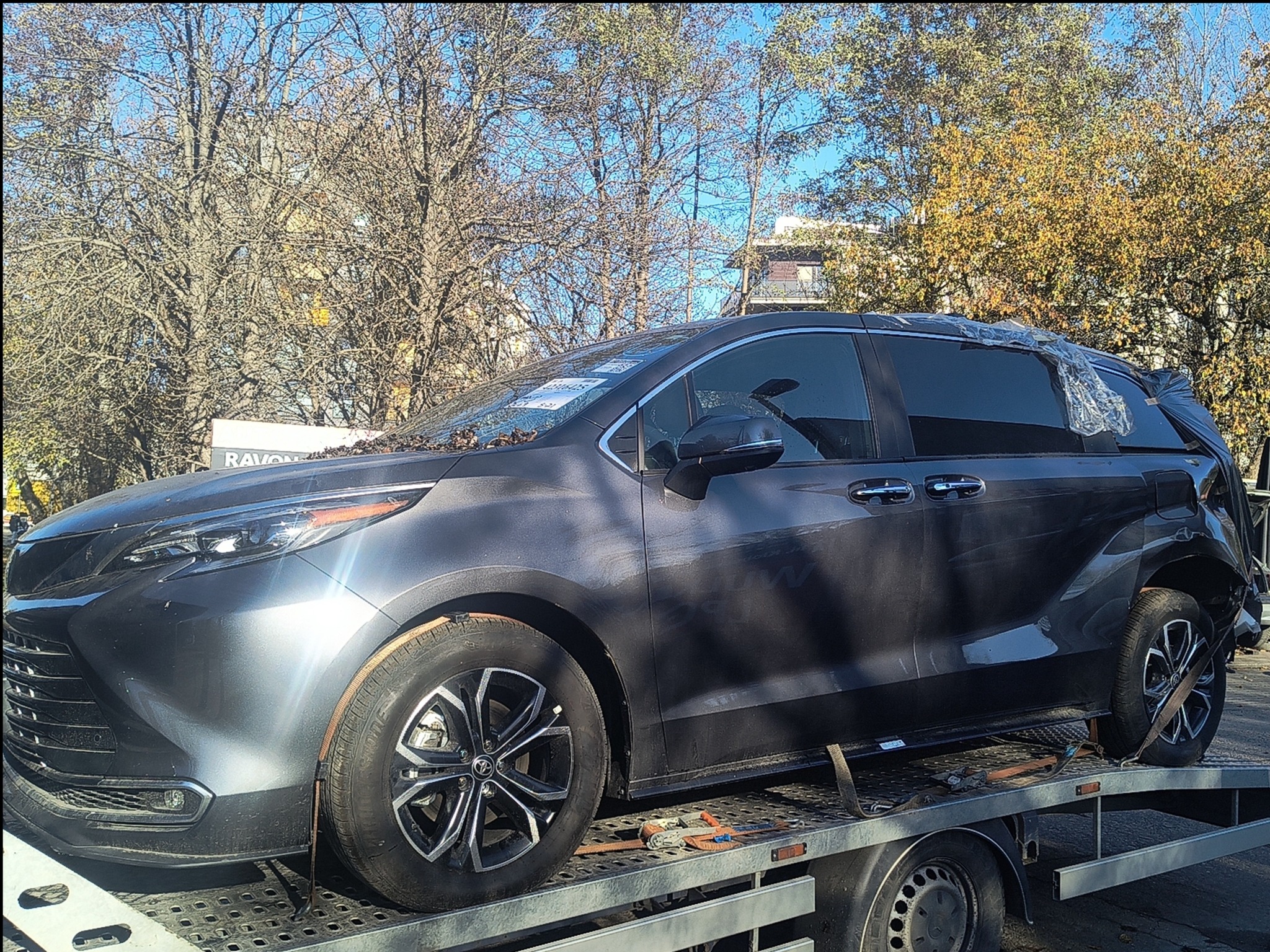 Uszkodzony, szary minivan na lawecie, zabezpieczony folią. Widoczne liście na karoserii, jesienne drzewa w tle. Detal: mocowanie kół pasami transportowymi.
