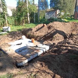 Dawid Gamułka dream's - Wykop z betonowym szambem w ziemi na tle zieleni i zabudowań. Widoczny sprzęt budowlany i rury. Ujęcie z góry, w słoneczny dzień.