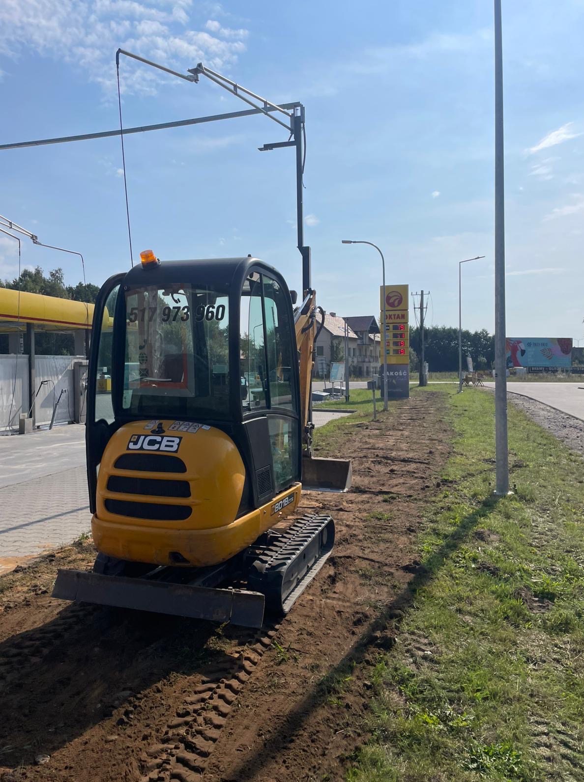 Żółta mini koparka JCB na ziemi obok chodnika i stacji benzynowej. Ślady opon w ziemi. Wykop pod instalacje.