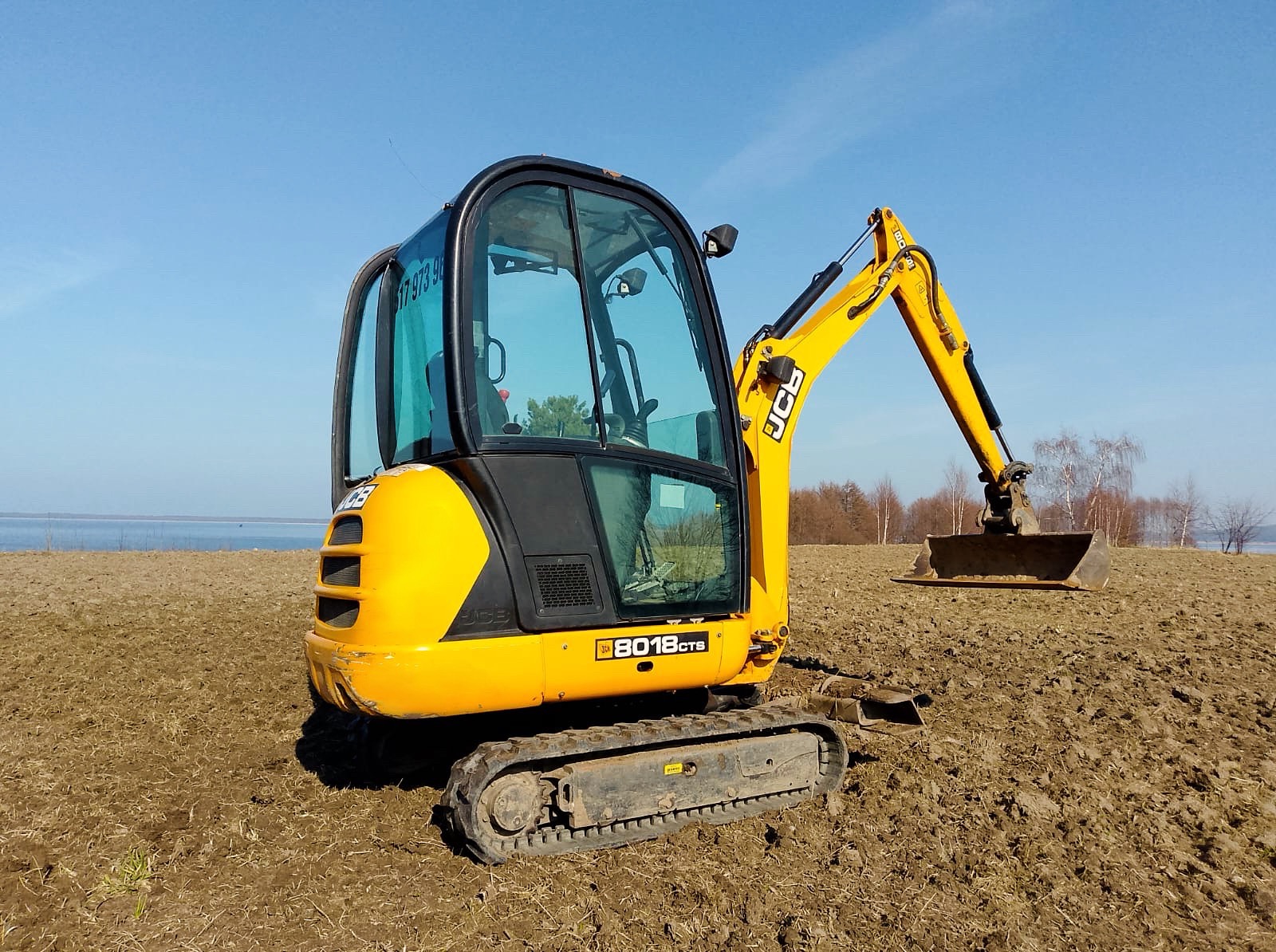 Żółta mini koparka JCB 8018CTS na polu z widokiem na jezioro w tle, błękitne niebo. Maszyna gotowa do pracy, łyżka lekko uniesiona.