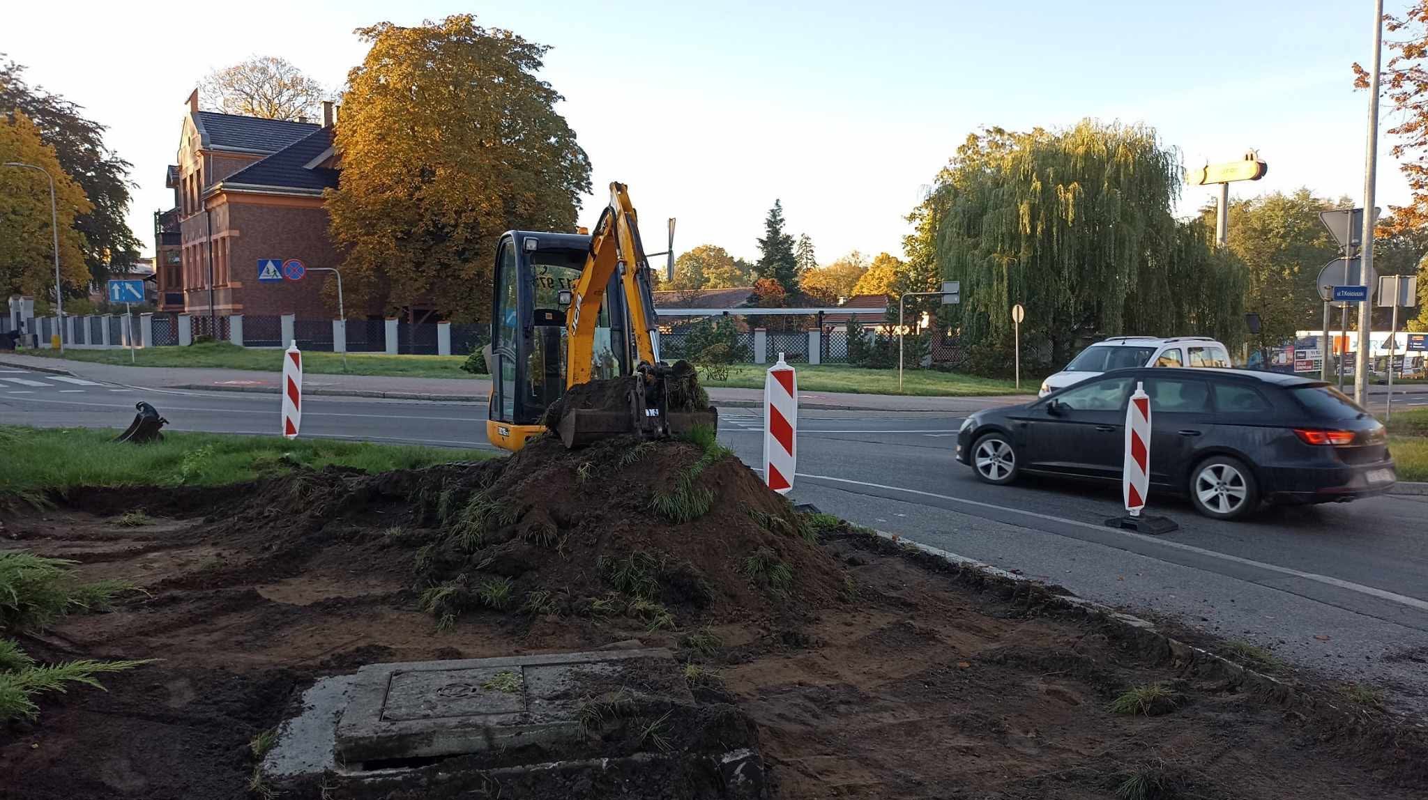 Żółta mini koparka na poboczu drogi, w tle budynek i przejeżdżające auto. Prace ziemne w przestrzeni miejskiej, wykop obok studzienki kanalizacyjnej.
