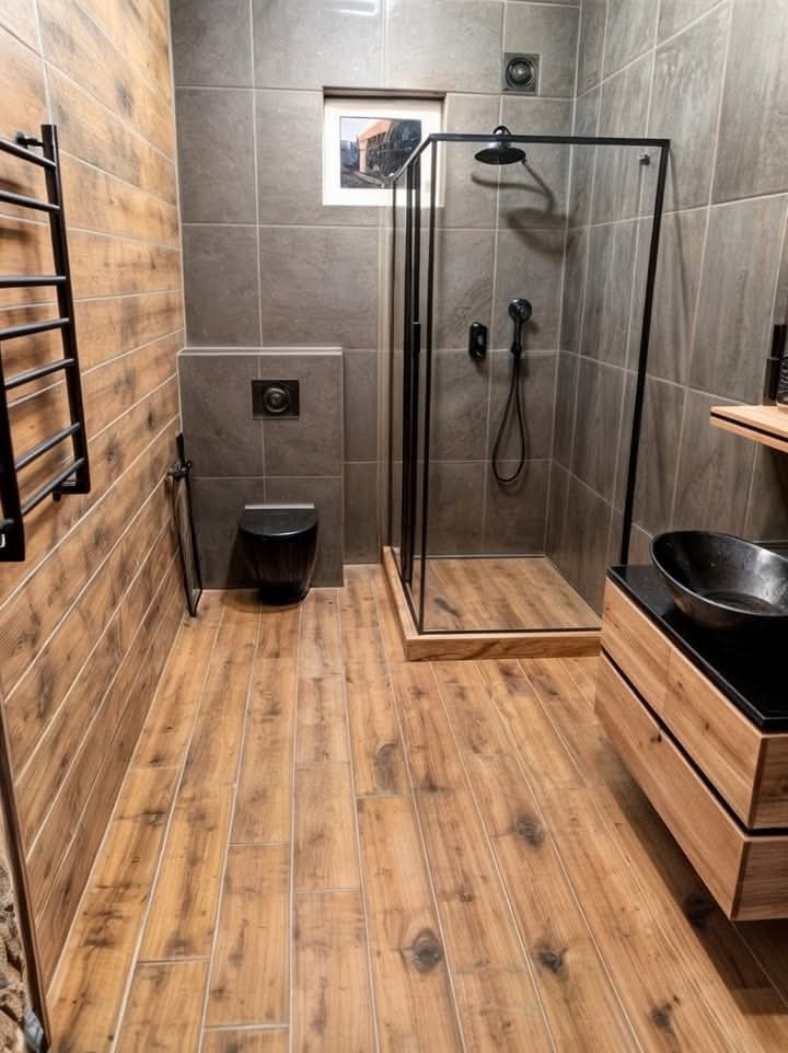 Nowoczesna łazienka z drewnopodobnymi płytkami na podłodze i ścianie, szara strefa prysznicowa z czarną kabiną, wisząca toaleta i umywalka nablatowa.