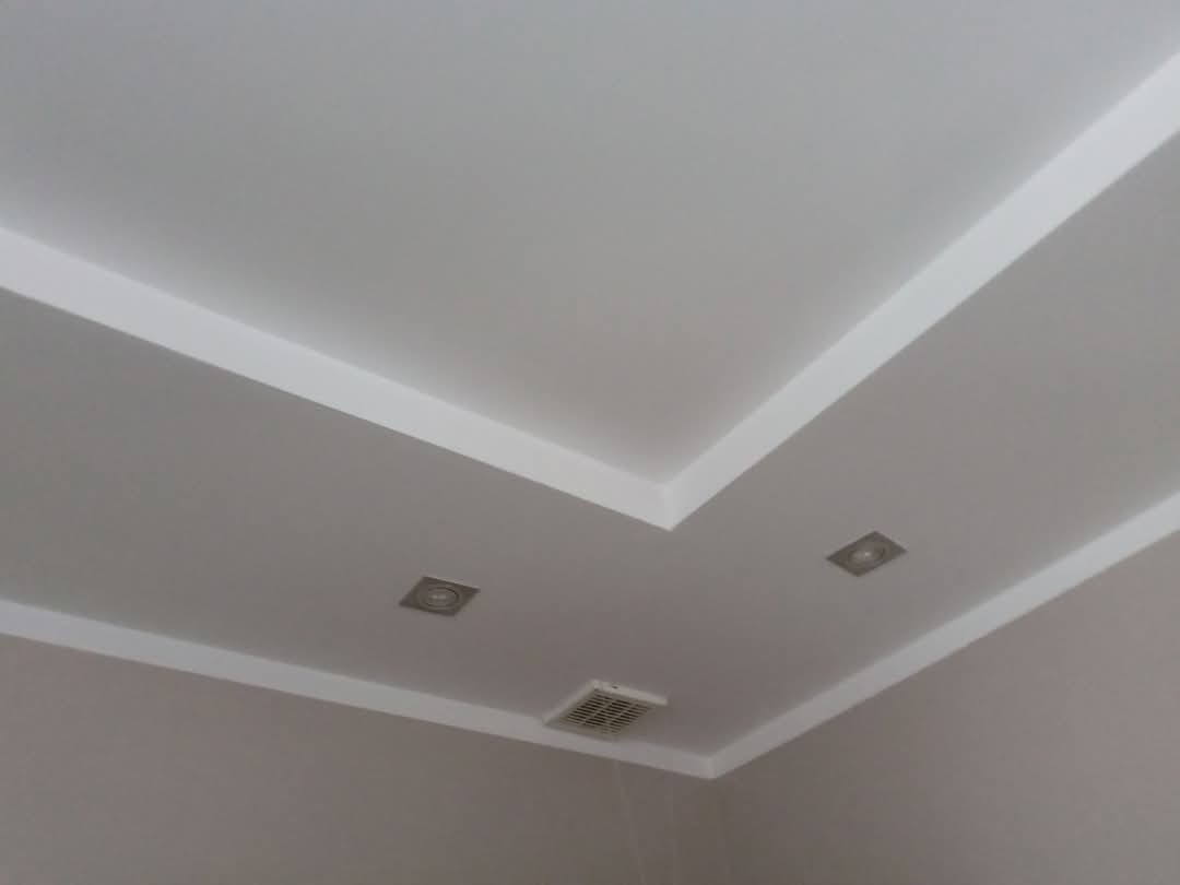 Biały sufit z listwami dekoracyjnymi, dwoma kwadratowymi lampami i kratką wentylacyjną. Minimalistyczny design, neutralne kolory, ukośny kadr.