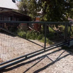 Nowoczesna, przesuwna brama wjazdowa z metalowej siatki panelowej na posesji z budynkiem w tle. Brama w kolorze grafitowym, zamontowana na betonowej podstawie.