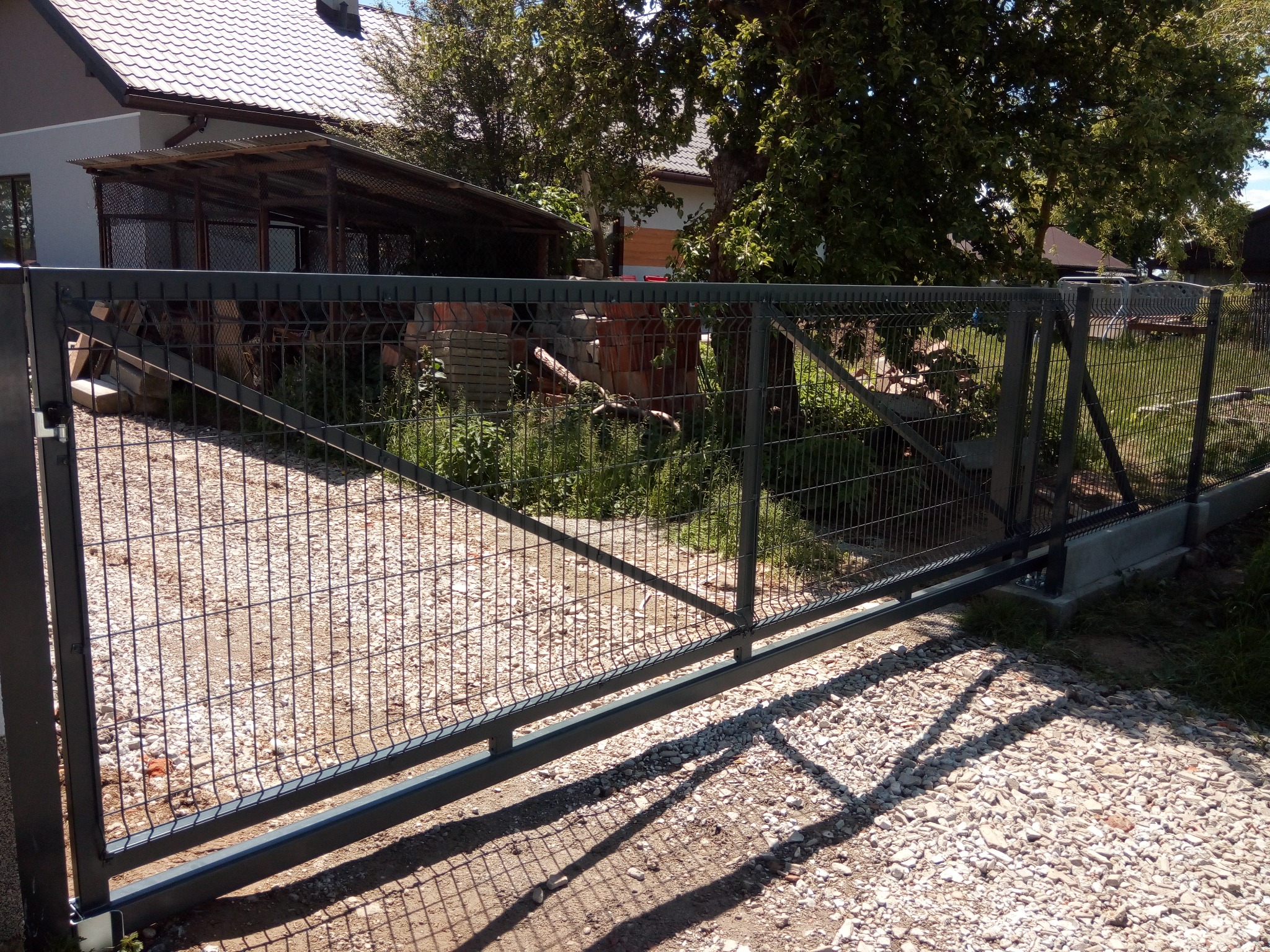 Nowoczesna, przesuwna brama wjazdowa z metalowej siatki panelowej na posesji z budynkiem w tle. Brama w kolorze grafitowym, zamontowana na betonowej podstawie.