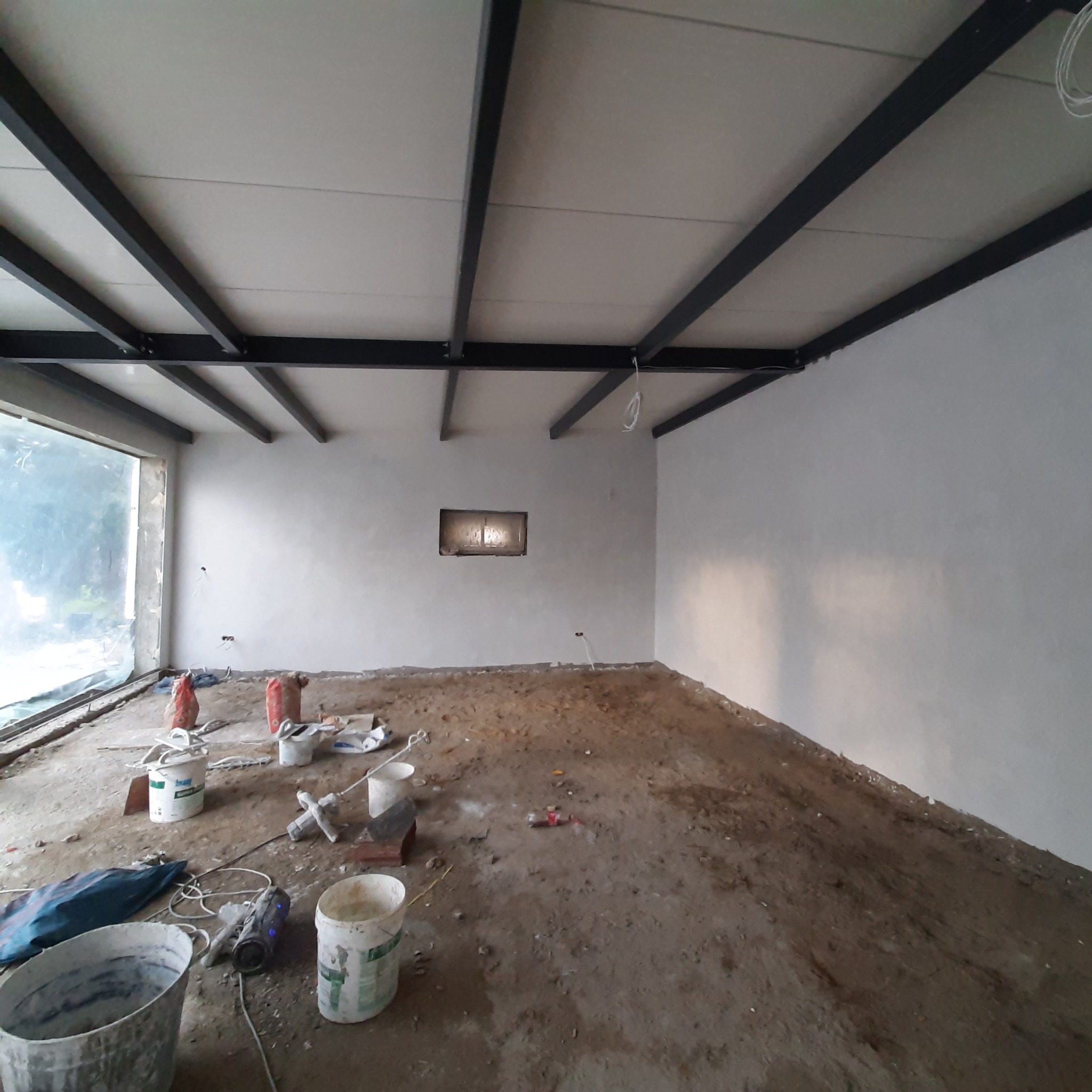 Wnętrze w trakcie tynkowania: świeżo otynkowane ściany, surowy strop z metalowymi belkami, widoczne narzędzia i materiały budowlane na zabrudzonej podłodze.