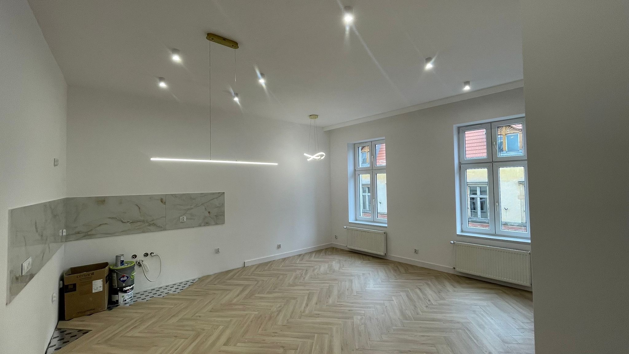 Jasny salon z aneksem kuchennym po remoncie: parkiet w jodełkę, marmurowe panele ścienne, nowoczesne oświetlenie. Widoczne okna i grzejniki. Świeżo po wykończeniu.