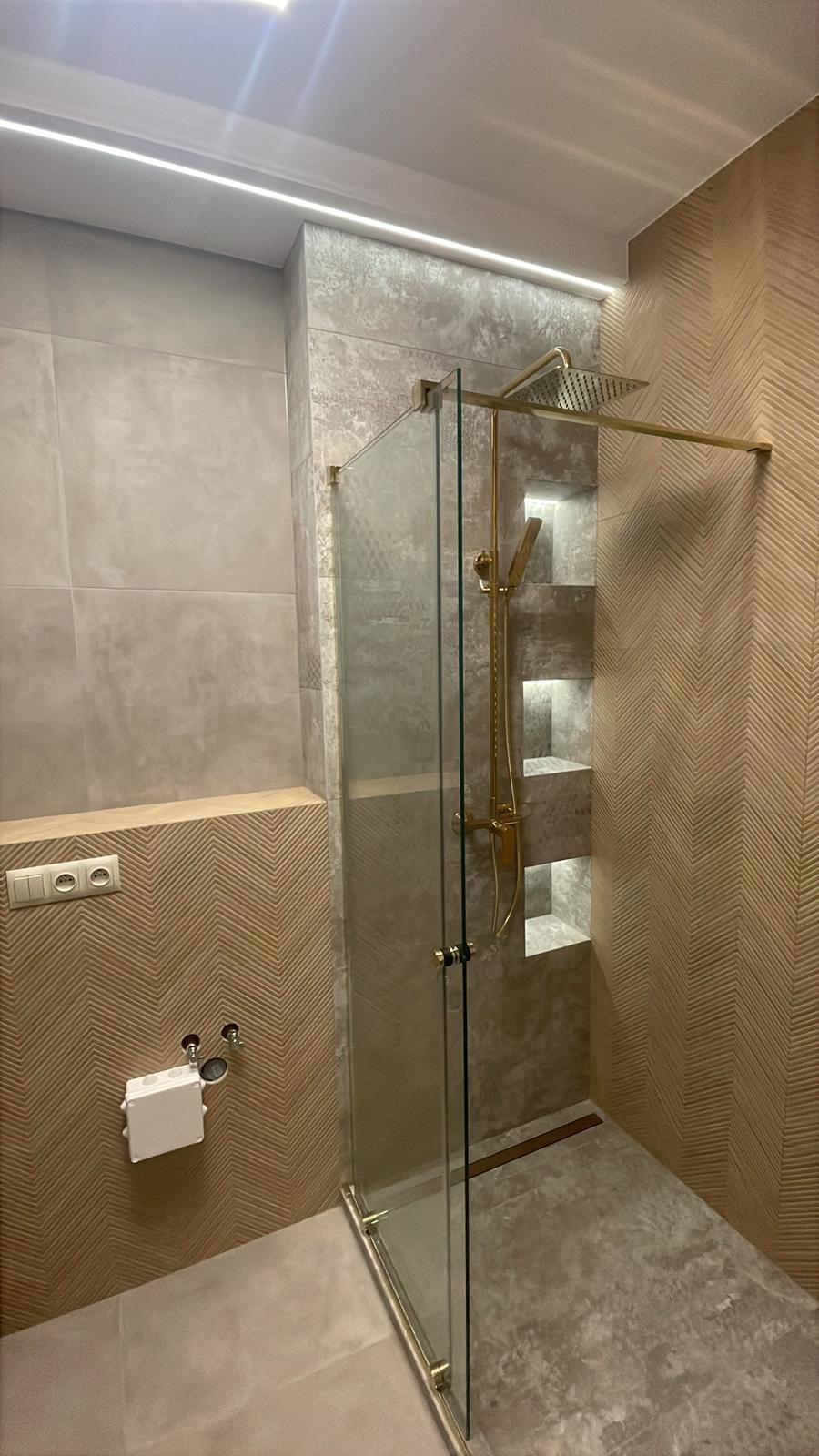 Nowoczesna łazienka z kabiną prysznicową typu walk-in, złotą armaturą i podświetlanymi niszami w ścianie. Dekoracyjne płytki w odcieniach beżu i szarości.