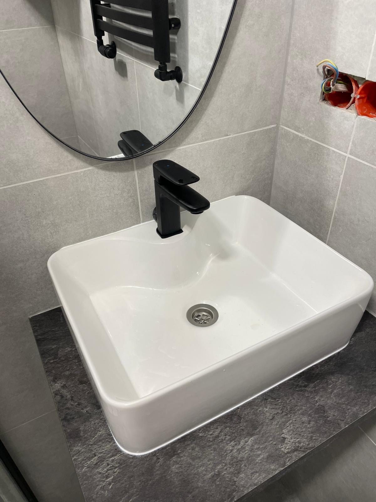 Biała, prostokątna umywalka z czarną baterią na blacie z ciemnego kamienia. W tle szare płytki, okrągłe lustro i widoczne przewody elektryczne.
