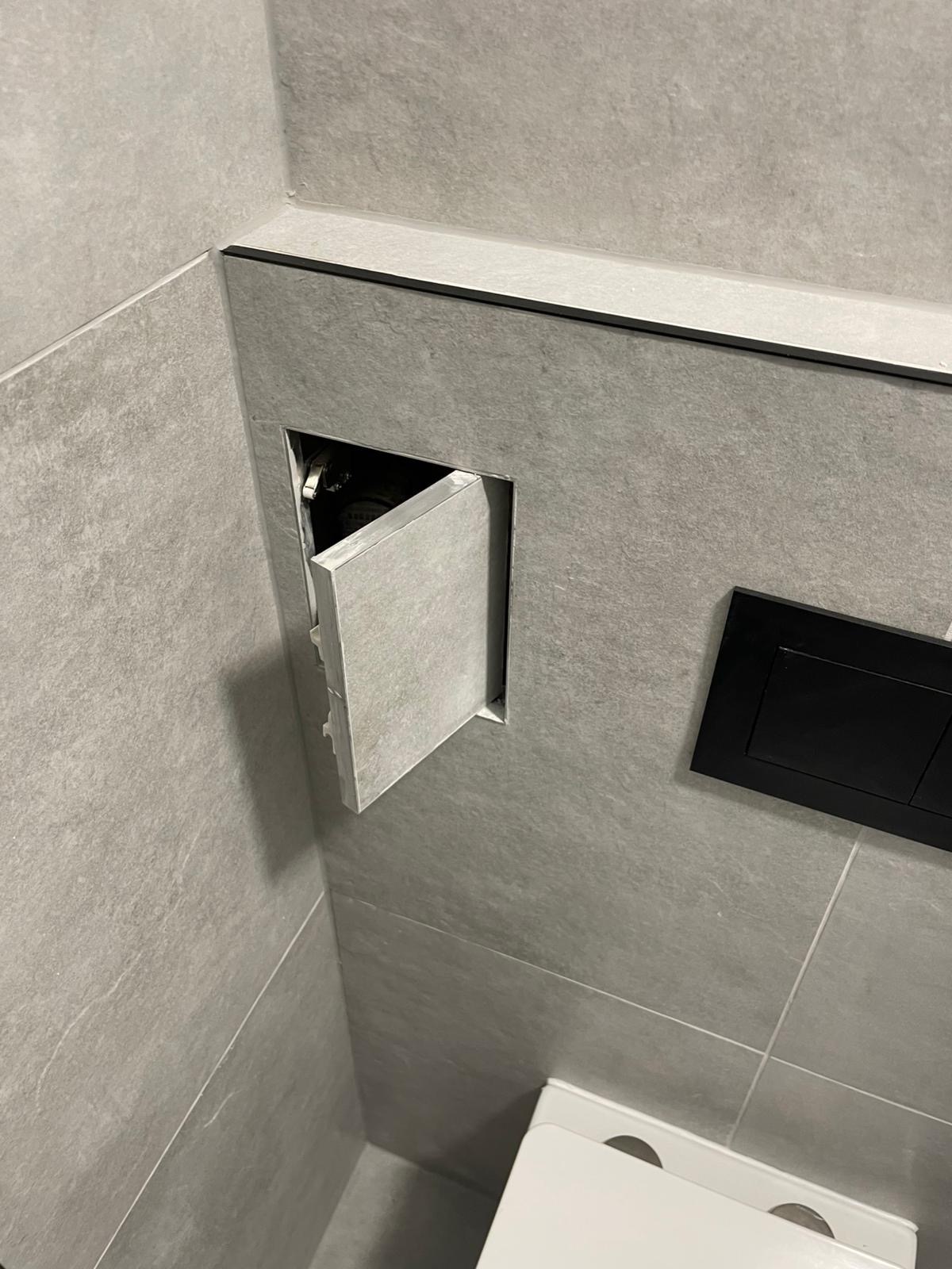 Szara toaleta z ukrytym panelem rewizyjnym, otwartym pod kątem. Widoczna czarna listwa i przycisk spłuczki. Minimalistyczny design, płytki imitujące kamień.