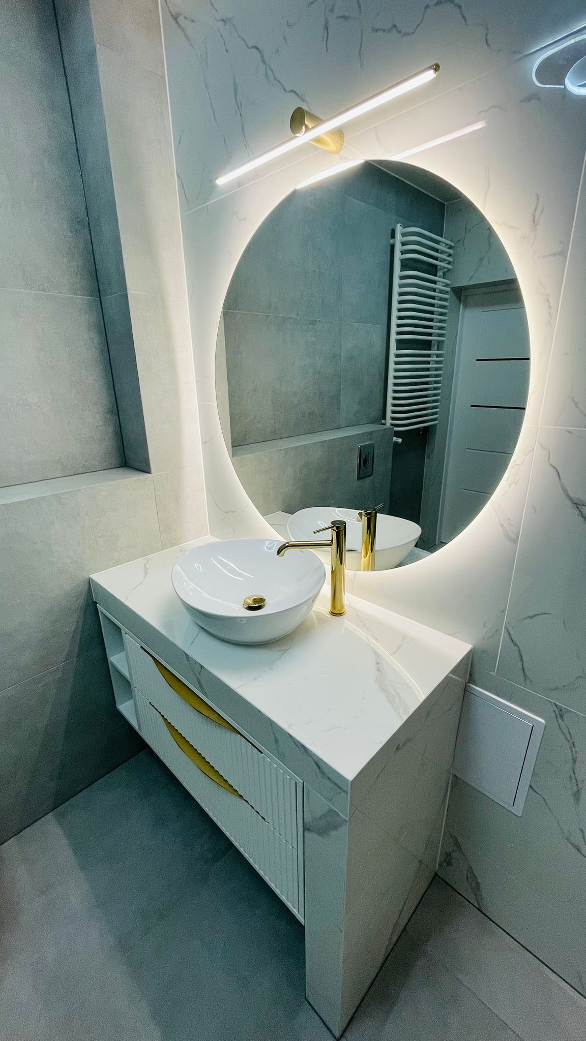 Nowoczesna łazienka z okrągłym lustrem LED, białą umywalką nablatową i szafką z marmurowym blatem oraz złotymi akcentami. Widoczne szare płytki imitujące marmur.