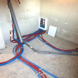 Łukasz Kozłowski - Rozdzielacz instalacji CO i wodnej z rurami PEX w kolorze niebieskim i czerwonym, zamontowany w surowym pomieszczeniu ze ścianami w trakcie wykończenia.