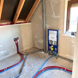 Łukasz Kozłowski - Instalacja wodno-kanalizacyjna w surowym wnętrzu poddasza: rury doprowadzające wodę (czerwone i niebieskie), szara rura odpływowa i stelaż podtynkowy Geberit.