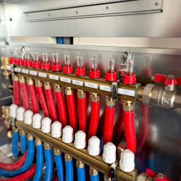 Łukasz Kozłowski - Rozdzielacz ogrzewania podłogowego z czerwonymi i niebieskimi rurami, zamontowany w metalowej szafce. Widoczne etykiety z nazwami pomieszczeń.