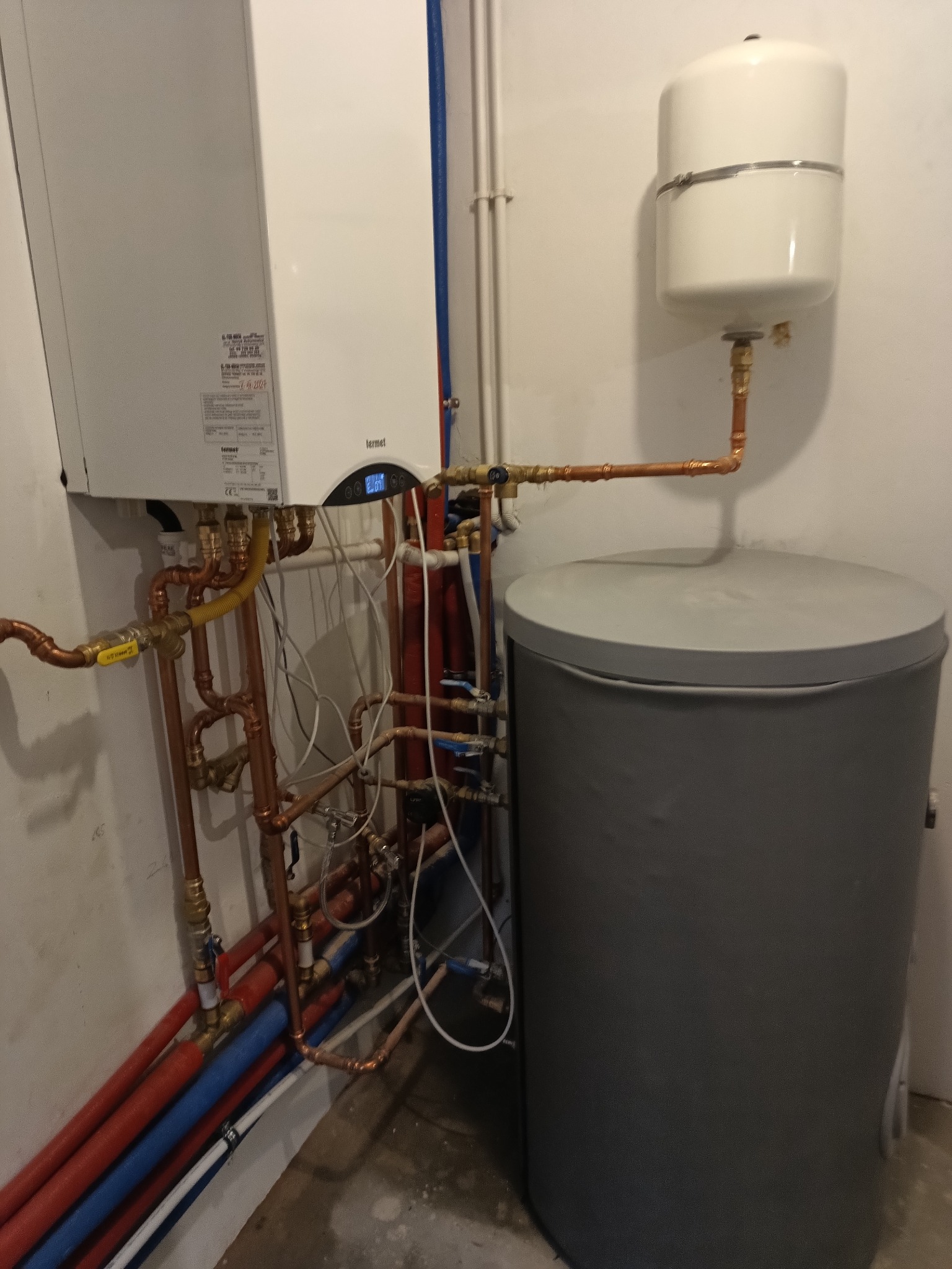 Kocioł gazowy Termet z podłączeniami miedzianymi i zbiornikiem akumulacyjnym. Widoczne rury w kolorach czerwonym i niebieskim. Instalacja w kotłowni.