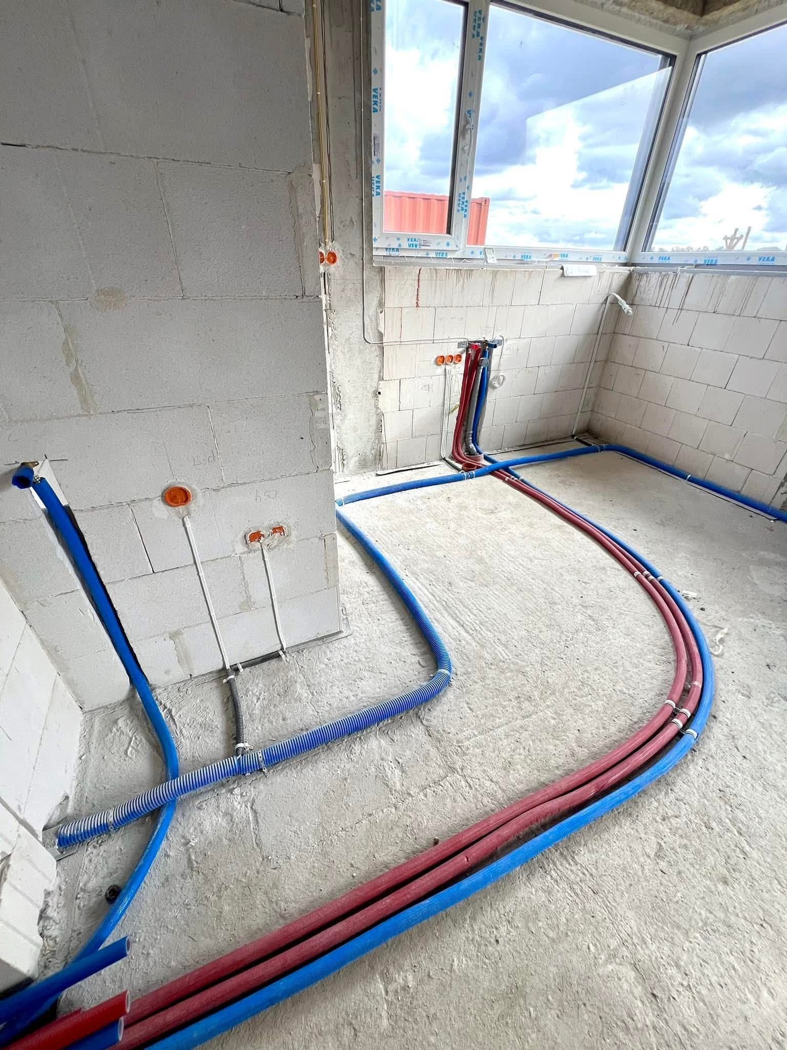 Wnętrze w trakcie instalacji: niebieskie i czerwone rury prowadzone po podłodze i ścianach z białego bloczka, widoczne okno z widokiem na niebo.