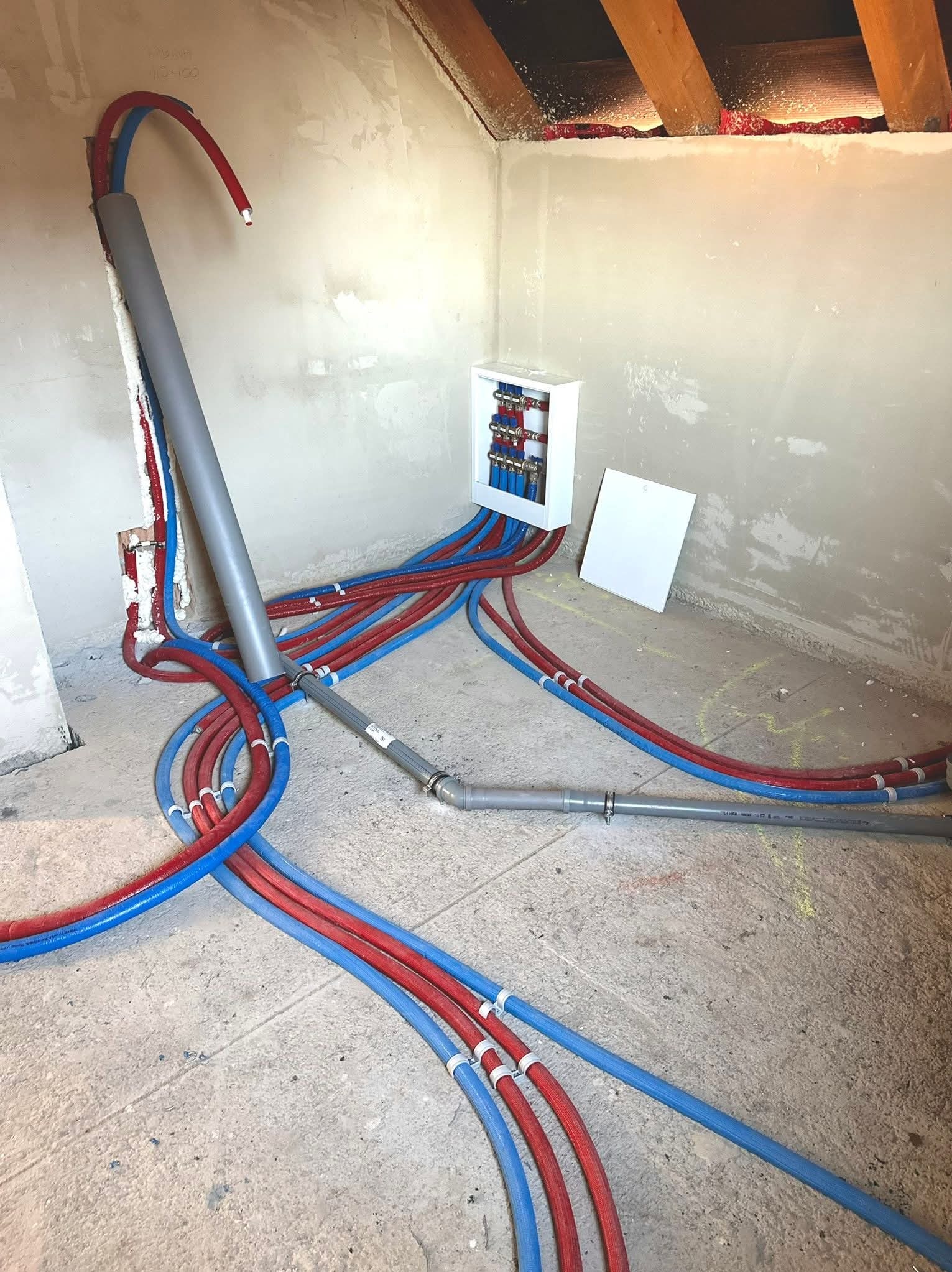 Rozdzielacz instalacji CO i wodnej z rurami PEX w kolorze niebieskim i czerwonym, zamontowany w surowym pomieszczeniu ze ścianami w trakcie wykończenia.