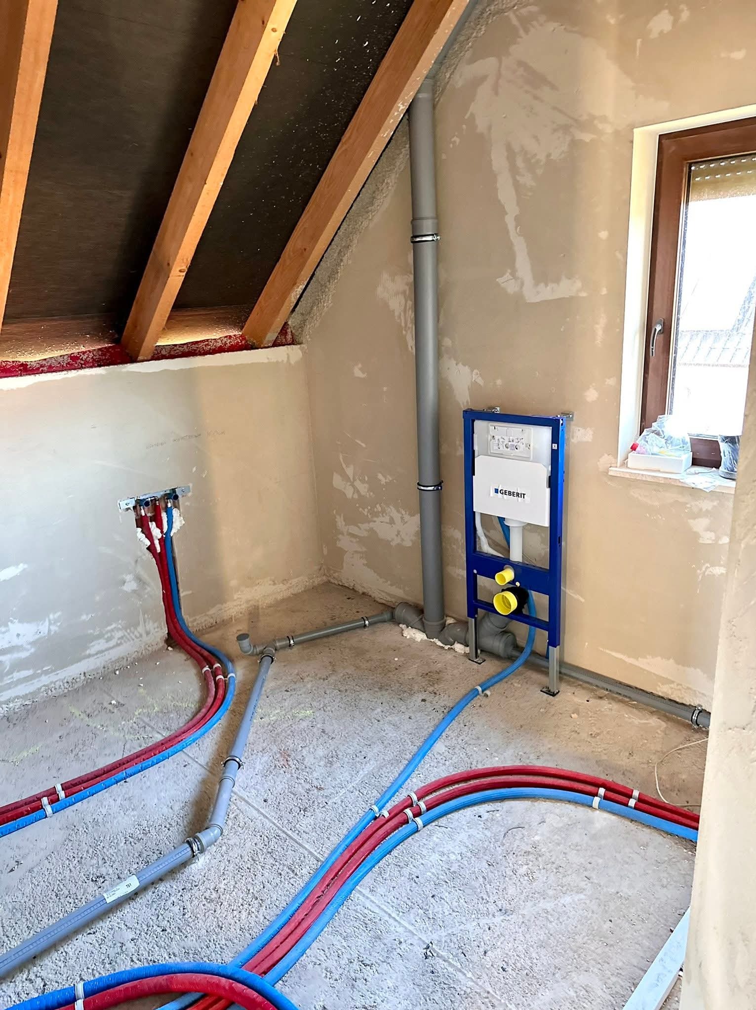 Instalacja wodno-kanalizacyjna w surowym wnętrzu poddasza: rury doprowadzające wodę (czerwone i niebieskie), szara rura odpływowa i stelaż podtynkowy Geberit.