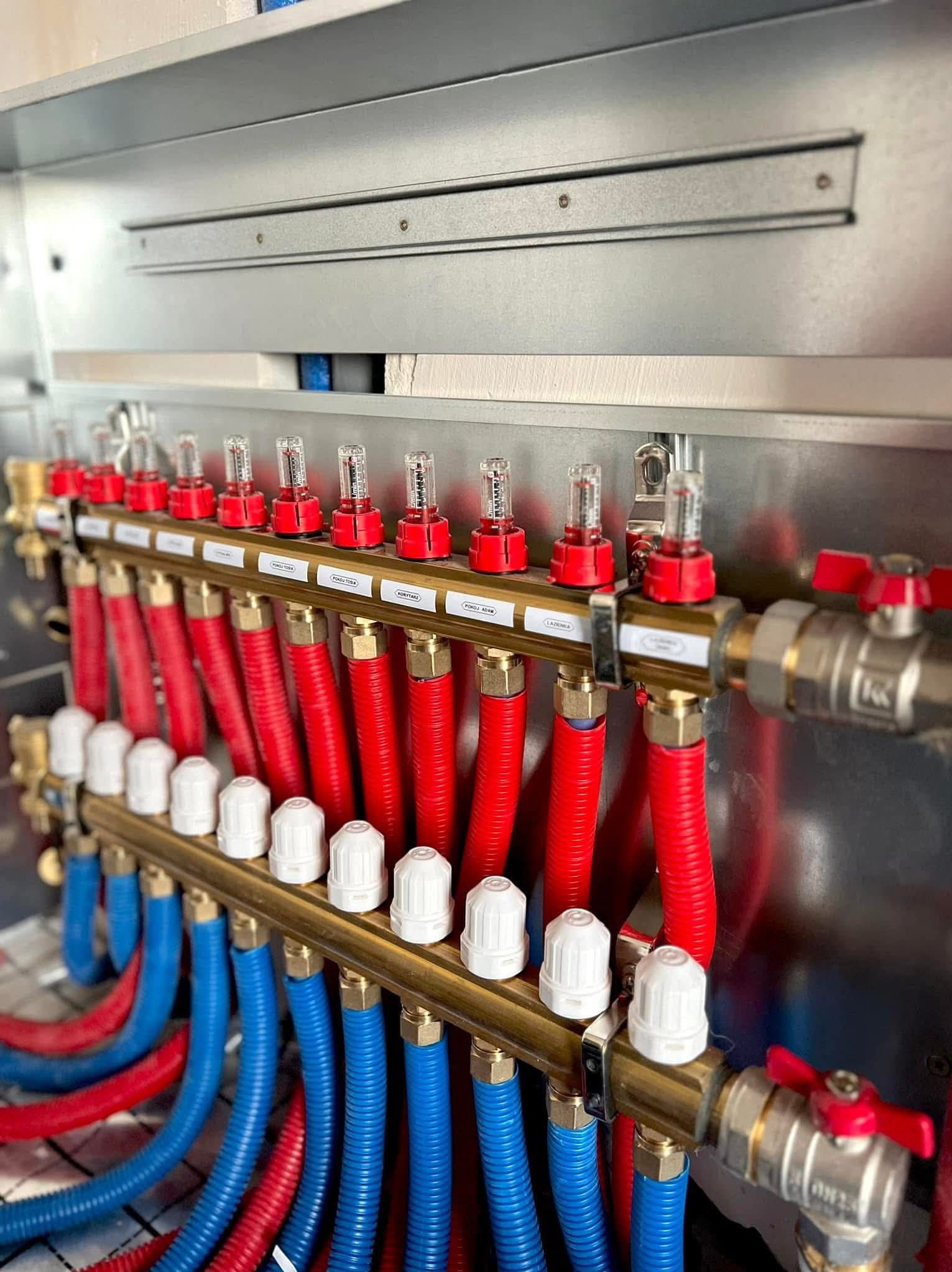 Rozdzielacz ogrzewania podłogowego z czerwonymi i niebieskimi rurami, zamontowany w metalowej szafce. Widoczne etykiety z nazwami pomieszczeń.