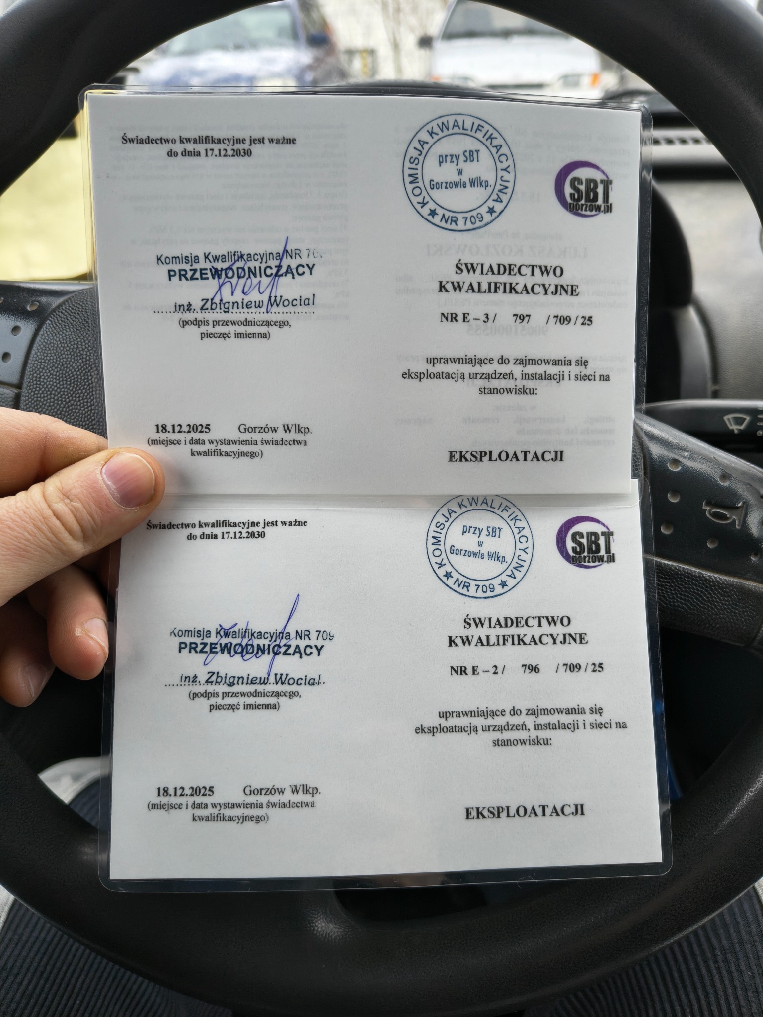 Świadectwo kwalifikacyjne z Gorzowa Wielkopolskiego, uprawniające do eksploatacji urządzeń, instalacji i sieci. Dokument trzymany w dłoni na tle kierownicy samochodu.