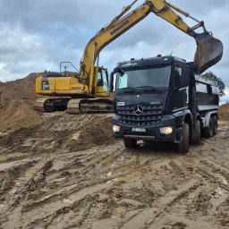 FIRMA KANADYS - Żółta koparka Komatsu ładuje piasek do czarnej ciężarówki Mercedes-Benz na błotnistym placu budowy, widok z przodu, pochmurne niebo w tle.