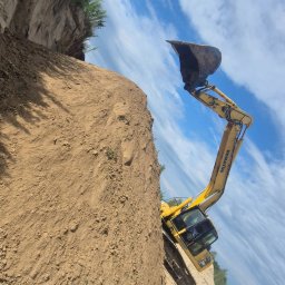 FIRMA KANADYS - Żółta koparka Komatsu na skraju wykopu, z widocznym niebem i roślinnością. Ujęcie z dołu podkreśla wysokość skarpy i zakres prac ziemnych.