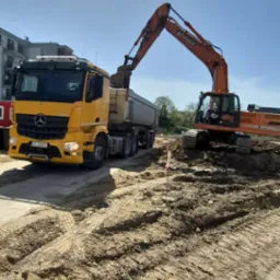 Pomarańczowa koparka Doosan ładuje ziemię do żółtej ciężarówki Mercedes-Benz na placu budowy w słoneczny dzień. Widoczne ślady opon na ziemi i budynek w tle.