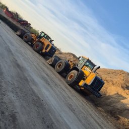 FIRMA KANADYS - Dwa żółte ładowarki kołowe JCB HL770-9 stojące na placu budowy, z hałdą piasku w tle i błękitnym niebem. Widoczne też inne maszyny budowlane.