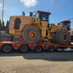 FIRMA KANADYS - Żółta ładowarka kołowa HL970 na czerwonej lawecie transportowej. Widoczne duże koła, kabina operatora i fragment otoczenia. Transport ciężkiego sprzętu budowlanego.