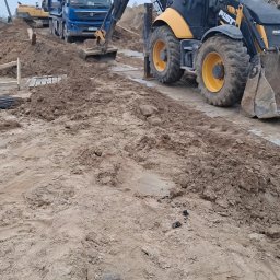 FIRMA KANADYS - Prace ziemne: koparka JCB, wywrotka Volvo i koparka gąsienicowa na placu budowy. W tle widoczne przygotowane elementy konstrukcyjne.