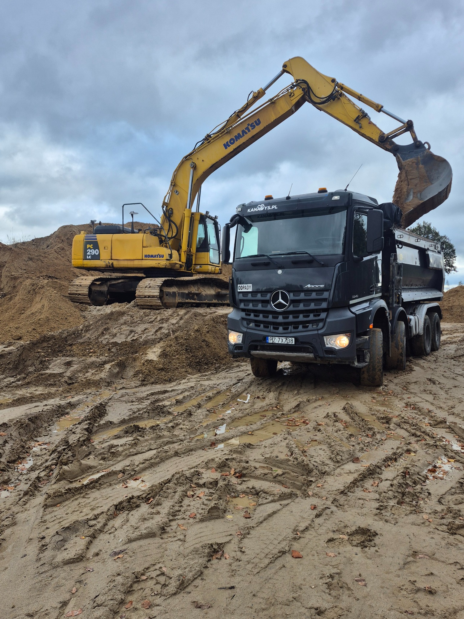 Żółta koparka Komatsu ładuje piasek do czarnej ciężarówki Mercedes-Benz na błotnistym placu budowy, widok z przodu, pochmurne niebo w tle.