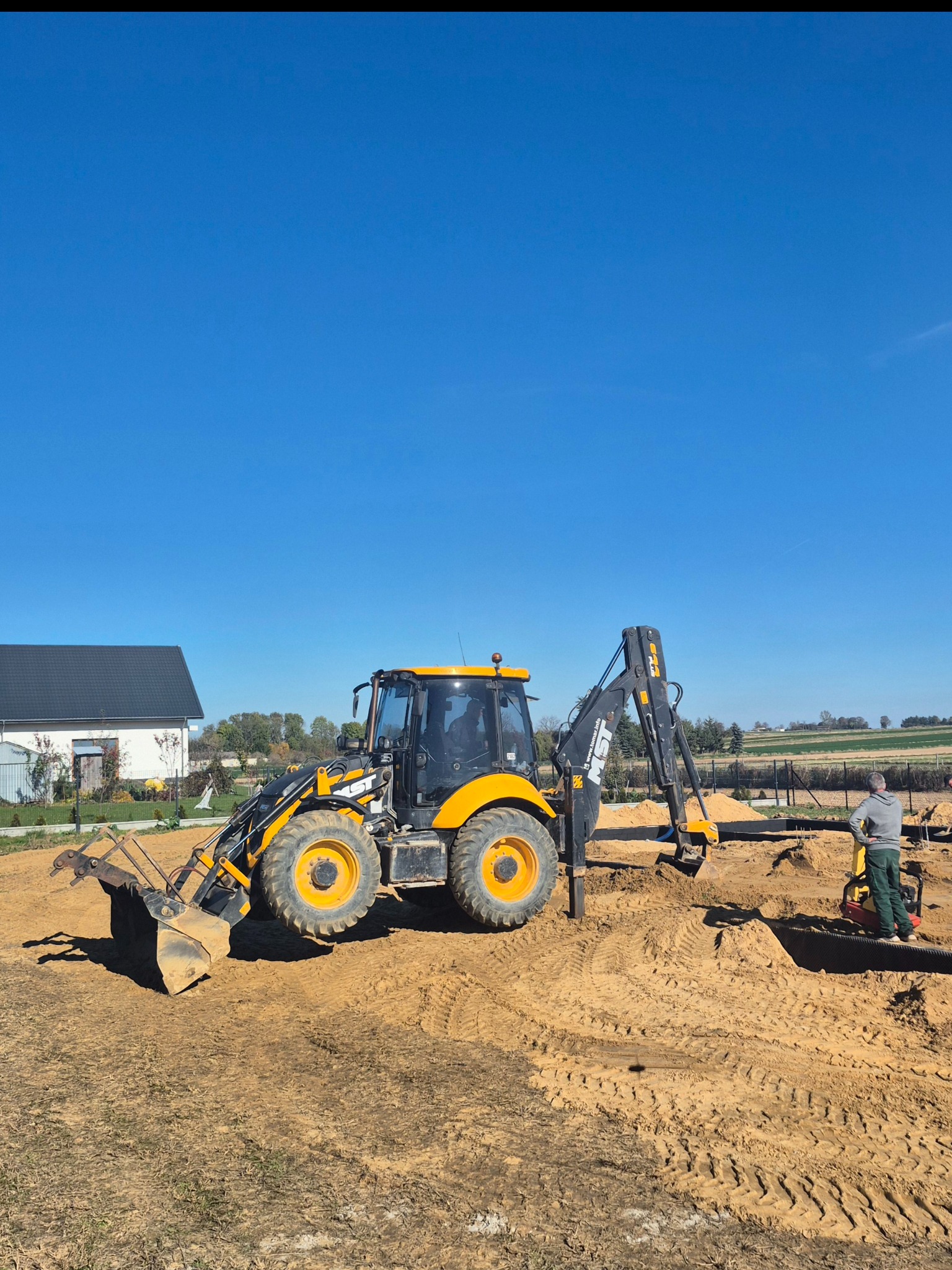 Żółto-czarna koparko-ładowarka JCB na placu budowy, z pracownikiem obsługującym zagęszczarkę w tle, przy wykopie fundamentów pod budynek na tle błękitnego nieba.